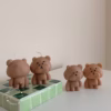 Three bear-shaped candles are placed on a white surface. Two of the candles are lit, with flames on top of their heads. The candles have round heads, small ears, and simple facial features. One candle is positioned on a green-tiled block, while the other two stand directly on the surface.