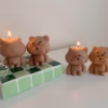 Three bear-shaped candles are placed on a white surface. Two of the candles are lit, with flames on top of their heads. The candles have round heads, small ears, and simple facial features. One candle is positioned on a green-tiled block, while the other two stand directly on the surface.