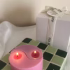 A pink yin-yang candle with two flames sits on a green and white tiled surface. A white gift box with a 