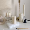 A minimal aesthetic display of decorative candles on a white surface. The arrangement includes a cube-shaped bubble candle, a tall embossed cylindrical candle, and two geometric block candles with angular patterns placed on a textured white base. Nearby are a few burnt matches, a black candle snuffer, and a brass candlestick holder. The scene has a soft, neutral color palette with beige and cream tones.