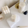A minimal aesthetic display of decorative candles on a white surface. The arrangement includes a cube-shaped bubble candle, a tall embossed cylindrical candle, and two geometric block candles with angular patterns placed on a textured white base. Nearby are a few burnt matches, a black candle snuffer, and a brass candlestick holder. The scene has a soft, neutral color palette with beige and cream tones.