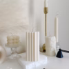 A minimal aesthetic display of decorative candles on a white surface. The arrangement includes a cube-shaped bubble candle, a tall embossed cylindrical candle, and two geometric block candles with angular patterns placed on a textured white base. Nearby are a few burnt matches, a black candle snuffer, and a brass candlestick holder. The scene has a soft, neutral color palette with beige and cream tones.