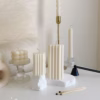 A minimal aesthetic display of decorative candles on a white surface. The arrangement includes a cube-shaped bubble candle, a tall embossed cylindrical candle, and two geometric block candles with angular patterns placed on a textured white base. Nearby are a few burnt matches, a black candle snuffer, and a brass candlestick holder. The scene has a soft, neutral color palette with beige and cream tones.
